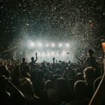 A large crowd gathers in the dark, facing a brightly lit stage with falling confetti.