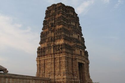 Ancient stone temple tower with intricate carvings set against a clear blue sky and greens.