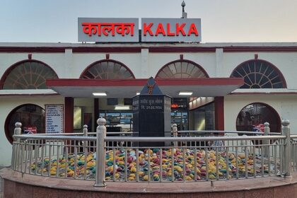 Kalka Railway Station in Haryana with passengers, parked trains, and signage, captured on a bright day with clear skies.