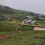 Foggy hillside view with scattered buildings, winding road, and lush green surroundings.