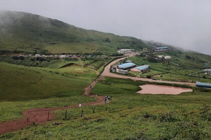 Foggy hillside view with scattered buildings, winding road, and lush green surroundings.