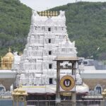 Towering white temple gopuram with golden domes surrounded by lush green hills behind.
