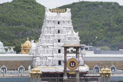 Towering white temple gopuram with golden domes surrounded by lush green hills behind.