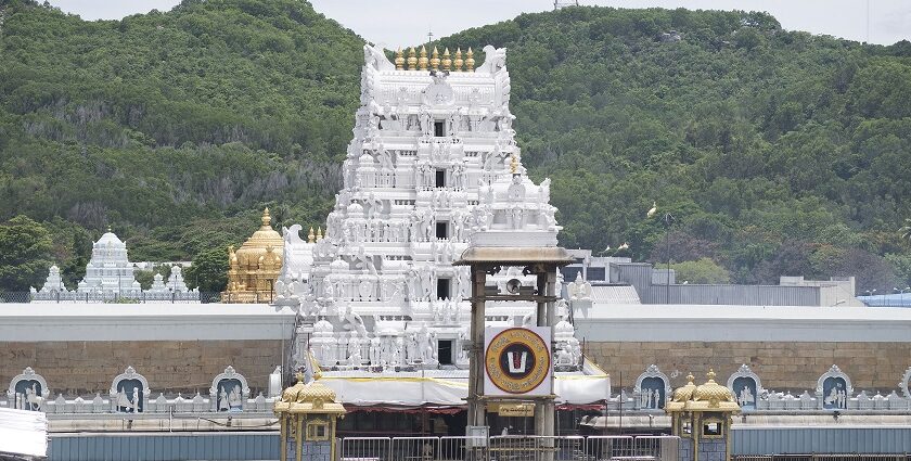 Towering white temple gopuram with golden domes surrounded by lush green hills behind.
