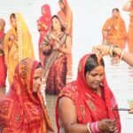 Women in traditional attire offering prayers during Chhath Puja at a riverbank.