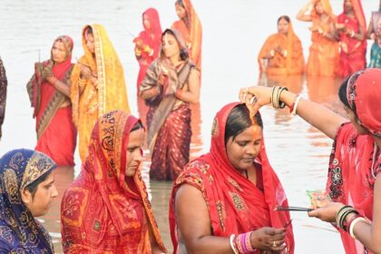Women in traditional attire offering prayers during Chhath Puja at a riverbank.