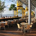 A waterfront outdoor cafe with many empty tables and chairs under a large, modern roof, one of the vegetarian restaurants in Singapore Little India.