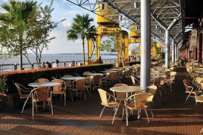 A waterfront outdoor cafe with many empty tables and chairs under a large, modern roof, one of the vegetarian restaurants in Singapore Little India.