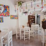 Bright restaurant with white wooden chairs, colourful wall art, hanging crafts and potted greens, one of the restaurants in Tezpur.