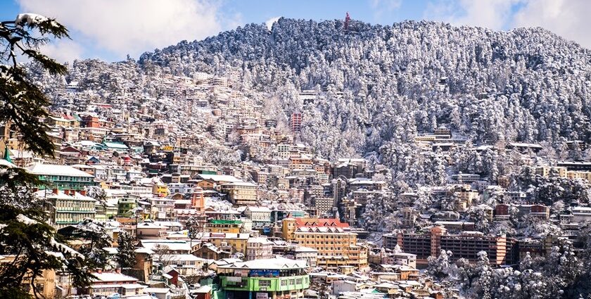 Scenic view of snowfall in Shimla with snow-covered roofs and mountain backdrop.