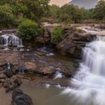 A huge river flows over rocky ledges at sunset, and there is a thick green forest all around it.