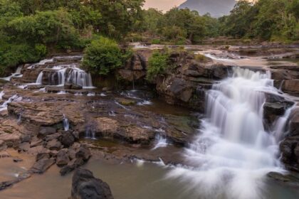 A huge river flows over rocky ledges at sunset, and there is a thick green forest all around it.