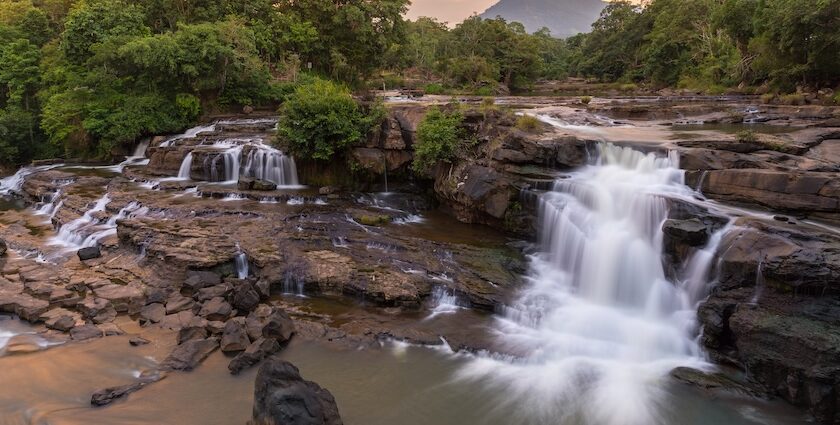 A huge river flows over rocky ledges at sunset, and there is a thick green forest all around it.